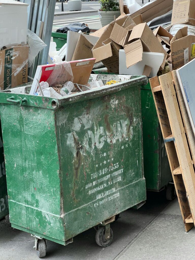 Green dumpster on city street filled with cardboard and debris, highlighting urban waste management.