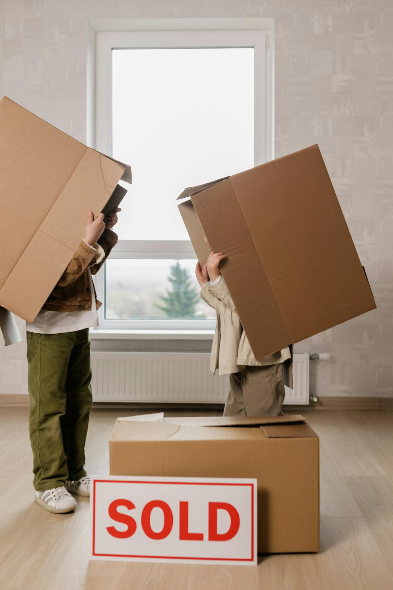 Two children playfully hide under boxes in their new home next to a sold sign.