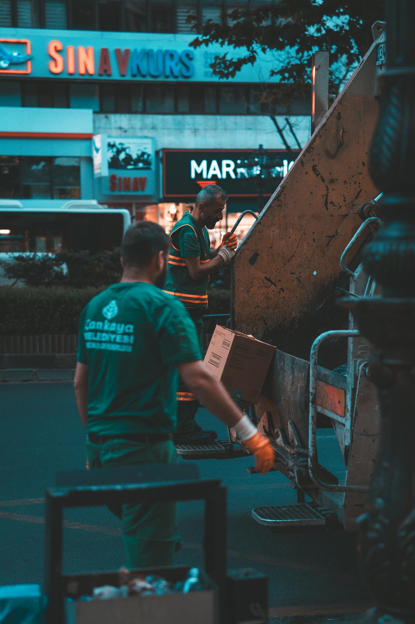 Two workers operate a garbage truck in a city street at night, wearing uniforms.